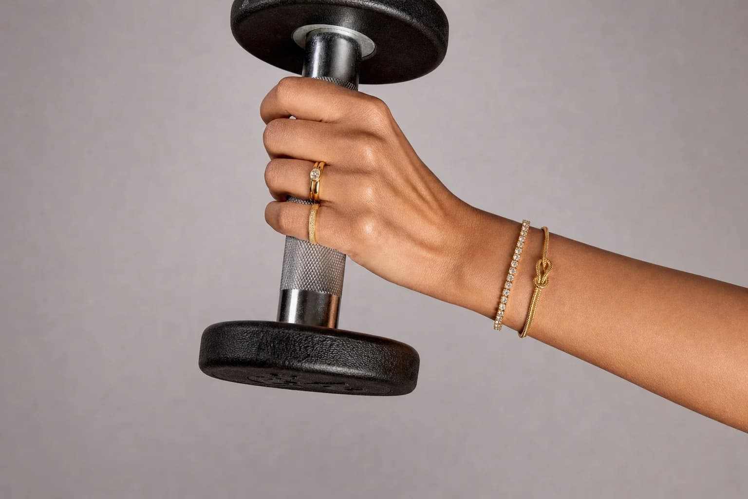 A hand gripping a dumbbell, the Crystal Tennis Bracelet and Knot Bangle at the wrist, the Luna Orbit and Stardust rings on the fingers