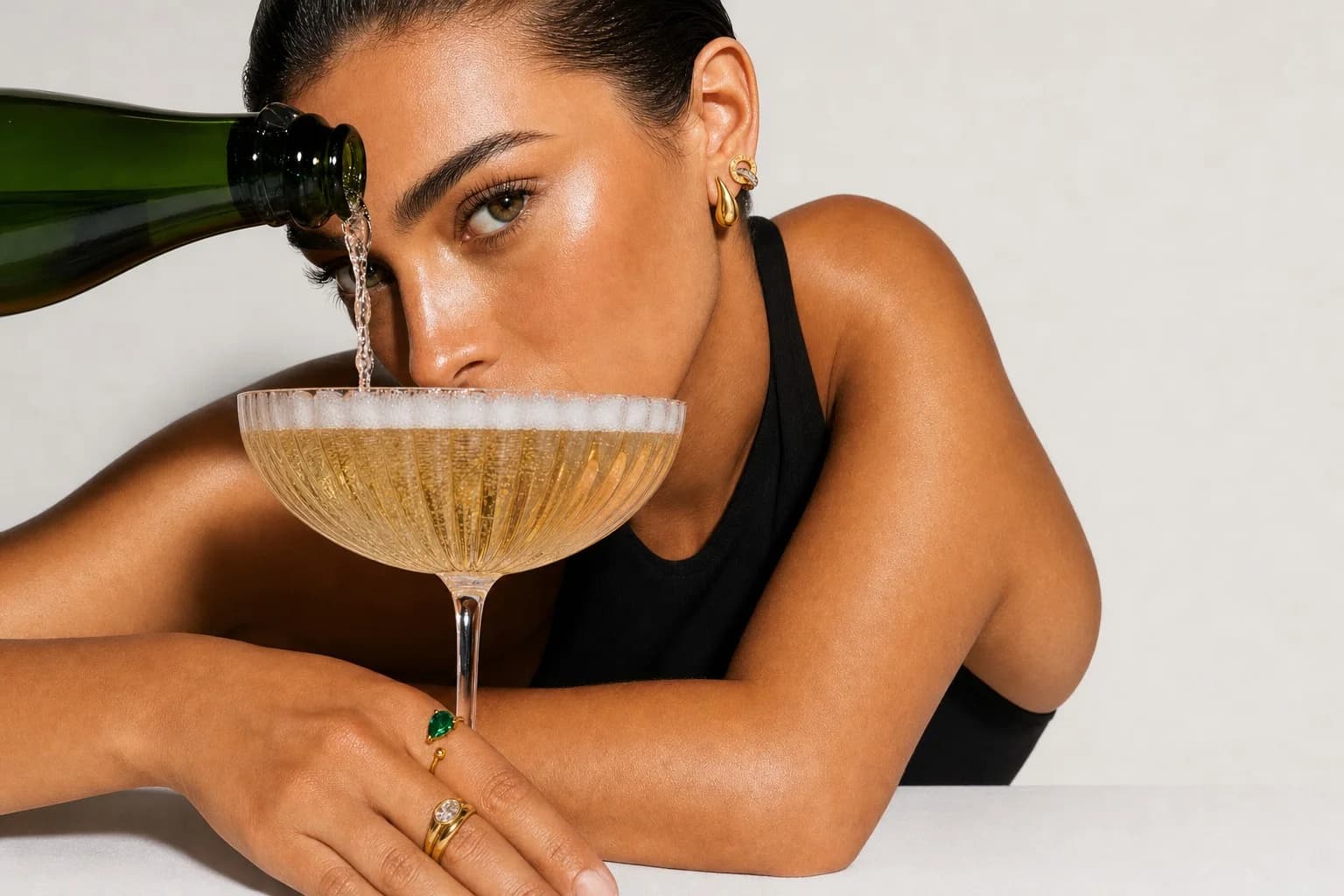 Champagne poured into a coupe at eye level, wearing the Chunky Teardrop Stud and Double Ring Stud, an emerald ring on the hand
