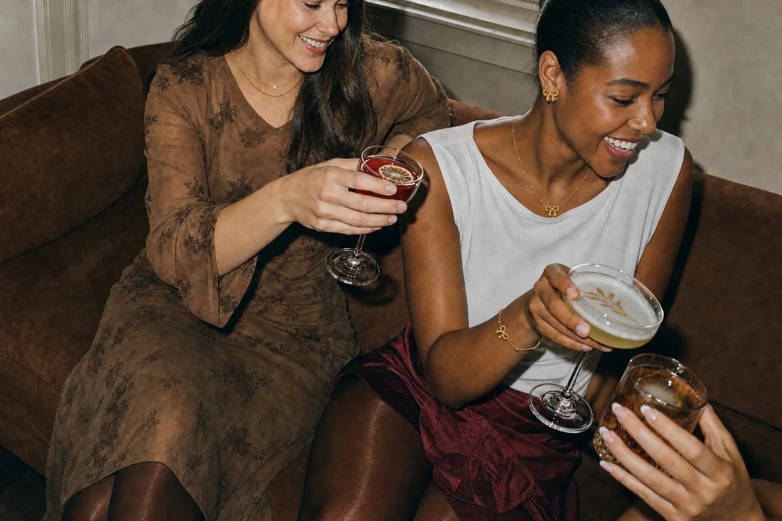 Two friends laughing over cocktails on a velvet sofa, one in the Serene Stack set, the other in the full Bow Knot set of studs, pendant and bracelet