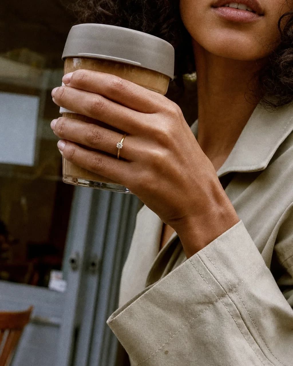 A hand around a coffee cup in a stone trench coat, wearing the Lumiere Solitaire Ring