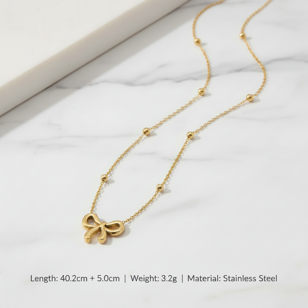 Gold necklace with bow pendant on a beige surface, showing measurements and material details.