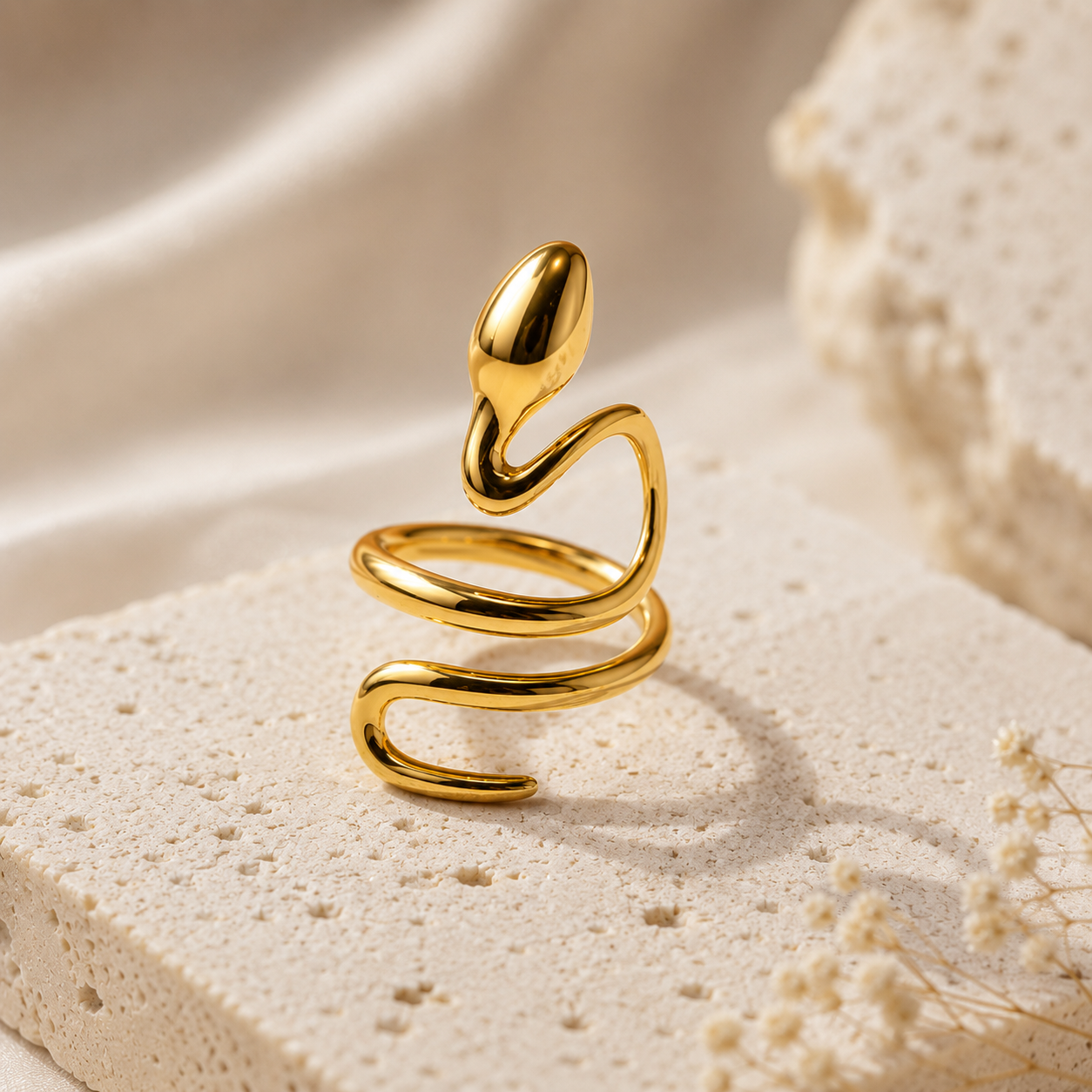 Gold snake,shaped ring on a textured beige surface with dried flowers.