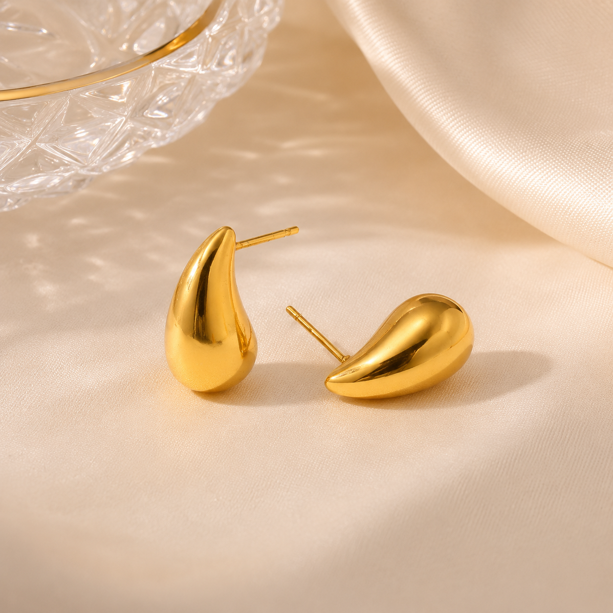 Gold earrings on a beige surface with a glass bowl in the background