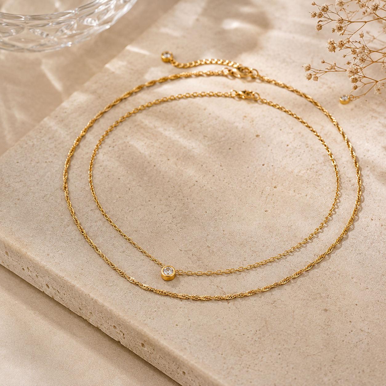 Two gold necklaces on a beige stone surface with a glass of water and dried flowers in the background.